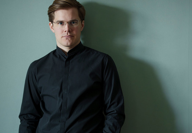 Man med glasögon ståendes mot en vägg. 